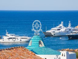 Blick über das türkiese Ziegeldach der Kirche Stella Maris auf große Motorjachten die im Hafen von Porto Cervo ankern Blick über das türkiese Ziegeldach der Kirche Stella Maris auf große Motorjachten die im Hafen von Porto Cervo ankern